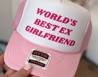 May include: A pink and white trucker hat with the text "WORLD'S BEST EX GIRLFRIEND" in pink. The hat features a foam front and a mesh back. The hat has a snap closure and a terry cloth sweatband.