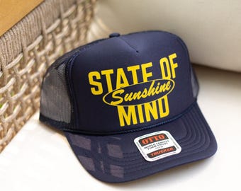 May include: Navy blue trucker hat with the text "STATE OF Sunshine MIND" in yellow. The hat has a mesh back and a curved brim. A white tag with black text is on the front.