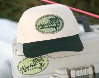 May include: A cream-colored baseball cap with a dark green brim and an oval patch featuring a fish graphic and the text Hookin AIN'T EASY. The cap sits on a light gray and dark green tackle box adorned with matching stickers of a fish.