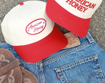 May include: Two white and red baseball caps with the text "American Honey" embroidered on them. One cap has the text in a script font and the other has the text in a block font.
