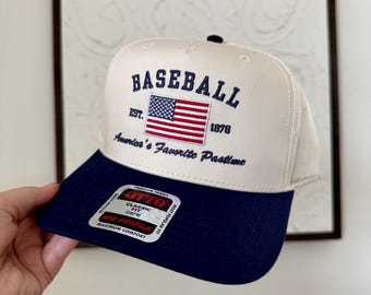 May include: A baseball cap with a cream-colored crown and a navy blue brim. The cap features the word "BASEBALL" above an American flag graphic, with "America's Favorite Pastime" below. The cap is a classic OSFM fit.