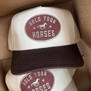 May include: White and brown baseball caps with the text "HOLD YOUR HORSES" in a maroon oval. The caps have a curved brim and a structured front panel. The design includes a horse graphic and star accents.