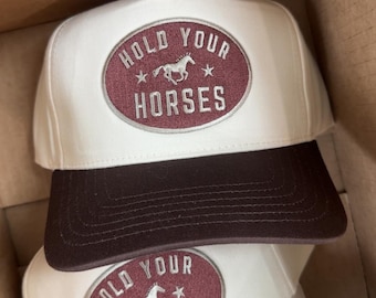 May include: White and brown baseball caps with the text "HOLD YOUR HORSES" in a maroon oval. The caps have a curved brim and a structured front panel. The design includes a horse graphic and star accents.