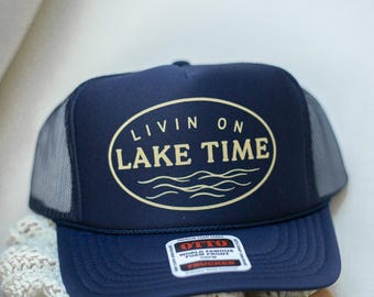 May include: Navy blue trucker hat with a mesh back and a curved brim. The hat features the text "LIVIN ON LAKE TIME" in an oval design. A white tag is visible on the front.