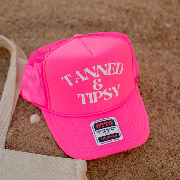 May include: Bright pink trucker hat with the words "TANNED & TIPSY" in white block letters. The hat is on a sandy surface, with a canvas tote bag partially visible in the lower left corner. A product tag is attached.