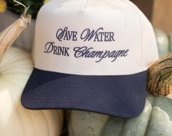 May include: Cream-colored baseball cap with a navy blue brim. The cap features the embroidered phrase "Save Water Drink Champagne" in navy blue script. The hat is displayed on a background of pumpkins and flowers.