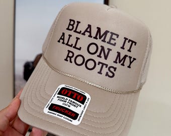 May include: A beige trucker hat with a black mesh back. The front of the hat has the text "BLAME IT ALL ON MY ROOTS" in black lettering. The hat has a black and red label that says "OTTO WORLD FAMOUS FOAM FRONT OSFM TRUCKER DURABLE SNAP CLOSURE".
