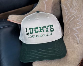 May include: A baseball cap with the text "LUCKY'S COUNTRY CLUB" in green lettering, featuring a white front panel and a dark green brim. The cap rests on a pair of tan cowboy boots.