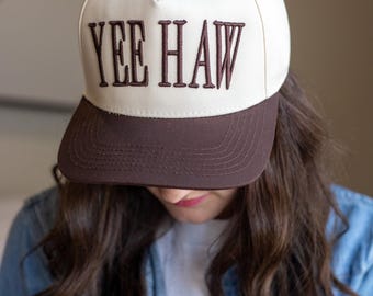 May include: A white and brown baseball cap with the text "YEE HAW" embroidered in brown. The cap has a curved brim and a button on top. The person in the image is wearing a denim jacket.