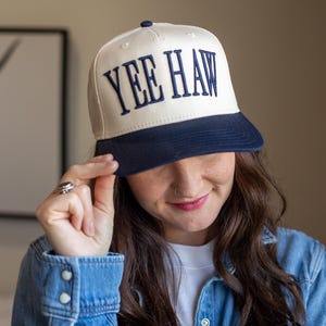 Puede incluir: Gorra de béisbol color crema con visera azul marino y el texto bordado "YEE HAW" en azul marino. La gorra tiene un diseño clásico de gorra de camionero.