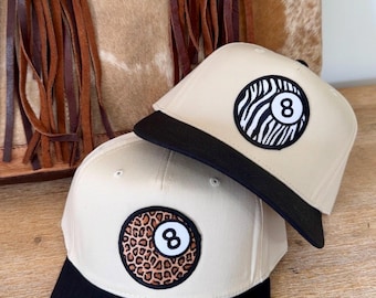 May include: Two beige baseball caps with black brims. Each cap features a circular patch with a white number 8. One patch has a zebra print, the other a leopard print. A leather bag with fringe is in the background.