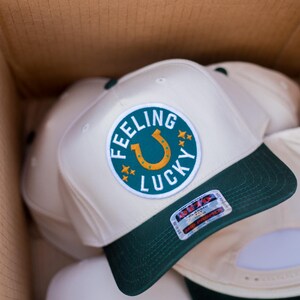May include: A stack of white baseball caps with green brims. The top cap has a green and gold embroidered patch that says "Feeling Lucky" with a horseshoe in the center.