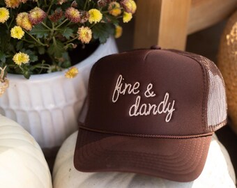 May include: A brown trucker hat with the embroidered phrase "fine & dandy" in white script. The hat has a mesh back and a curved brim. The hat is resting on a white pumpkin, with a white ceramic pot of yellow flowers in the background.