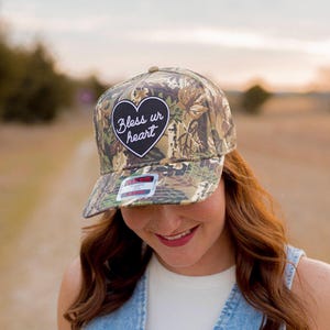 May include: A camouflage baseball cap with a black heart patch that says "Bless ur heart" in white lettering.