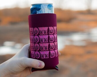 May include: A maroon can cooler with the words "Howdy" repeated in pink lettering. The cooler is holding a blue and white can.