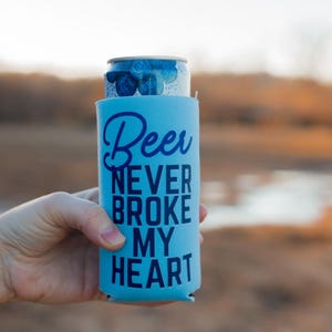 Puede incluir: Un enfriador de latas azul con el texto "Beer never broke my heart" en letras azul oscuro.