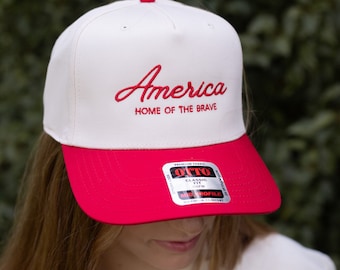 May include: White baseball cap with a red brim. The front of the cap has red embroidered text that reads "America Home of the Brave".