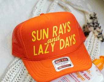 May include: Orange trucker hat with the text "SUN RAYS and LAZY DAYS" in yellow. A bottle of b.tan after-sun lotion is next to the hat. The hat has a mesh back and a foam front.