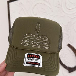 Könnte beinhalten: Eine olivgrüne Trucker-Cap mit Netzrücken und strukturierter Schaumstoff-Vorderseite. Auf der Vorderseite befindet sich ein weißes, gesticktes Kronenmotiv. Ein rechteckiger Aufkleber auf dem Schirm zeigt OTTO WORLD FAMOUS FOAM FRONT OSFM TRUCKER.