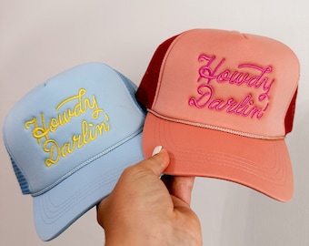 Puede incluir: Dos gorras de camionero con espalda de malla. Una gorra es azul claro con bordado amarillo que dice "Howdy Darlin'" y la otra gorra es melocotón con bordado rosa que dice "Howdy Darlin'"