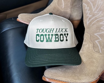 May include: A white and green baseball cap with the text "TOUGH LUCK COWBOY" in green lettering. The cap is paired with a pair of tan cowboy boots.