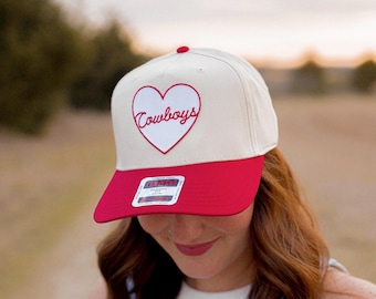 May include: A white and red baseball cap with a red heart embroidered on the front. The heart has the word "Cowboys" written inside it in red cursive.