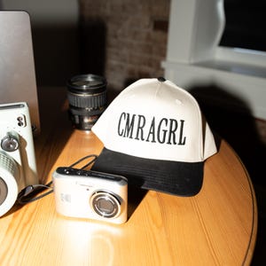 May include: A cream-colored baseball cap with a black brim and the text "CMRAGRL" in black block letters. A silver digital camera and a black camera lens are also on the wooden surface.
