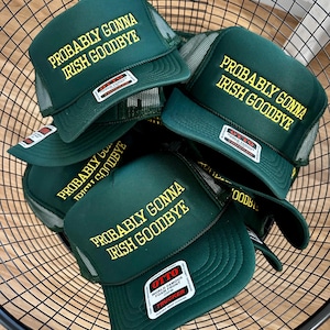 May include: A selection of dark green trucker caps, each embroidered with the yellow text "PROBABLY GONNA IRISH GOODBYE". The caps feature a mesh back and a curved peak. They are displayed in a black wire basket.