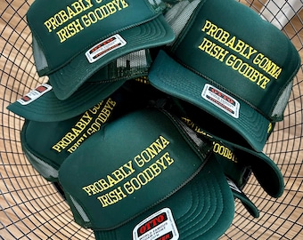 May include: A collection of dark green trucker hats with the phrase "PROBABLY GONNA IRISH GOODBYE" embroidered in yellow. The hats have a mesh back and a curved brim. The hats are in a black wire basket.