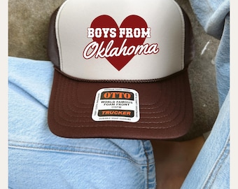 May include: A brown and white trucker hat with a red heart and the text "BOYS FROM Oklahoma" in white. The hat has a black mesh back and a brown bill. The hat has a label that says "OTTO WORLD FAMOUS FOAM FRONT OSFM TRUCKER DURABLE SNAP CLOSURE"