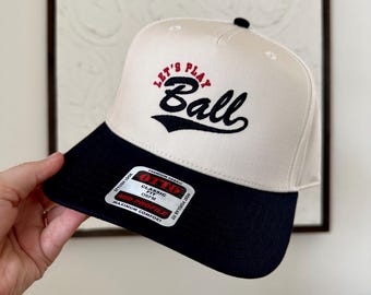 May include: A cream-colored baseball cap with a navy blue brim. The cap features the text "Let's Play Ball" in red and black script. The cap has a classic fit and is made with premium fabric.