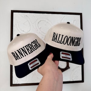 May include: Two baseball caps with cream-colored crowns and navy blue brims. One cap reads "BANNERGRL" and the other reads "BALLOONGRL" in bold black letters. Each cap has a black button on top and a label on the brim.