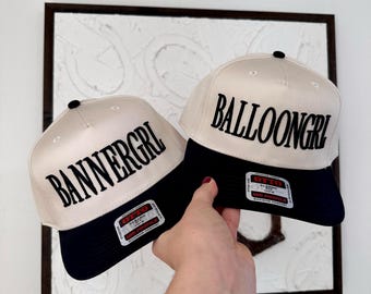 May include: Two baseball caps with cream-colored crowns and navy blue brims. One cap reads "BANNERGRL" and the other reads "BALLOONGRL" in bold black letters. Each cap has a black button on top and a label on the brim.