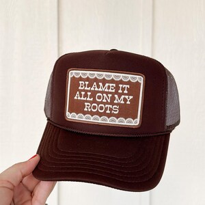 May include: A dark brown trucker hat featuring a rectangular patch with white lace-style trim. The patch contains the white text "BLAME IT ALL ON MY ROOTS" in a bold, sans-serif font. The hat has a solid front panel and a mesh back panel.
