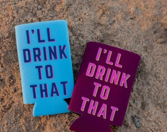 May include: Two blue and purple can coolers with the text "I'll drink to that" printed on them. The coolers are sitting on a sandy surface.