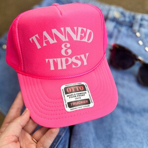 May include: A bright pink trucker hat with the words "TANNED & TIPSY" in white block letters. The hat has a mesh back and a curved brim. The hat also has a tag that says "OTTO" and "TRUCKER".