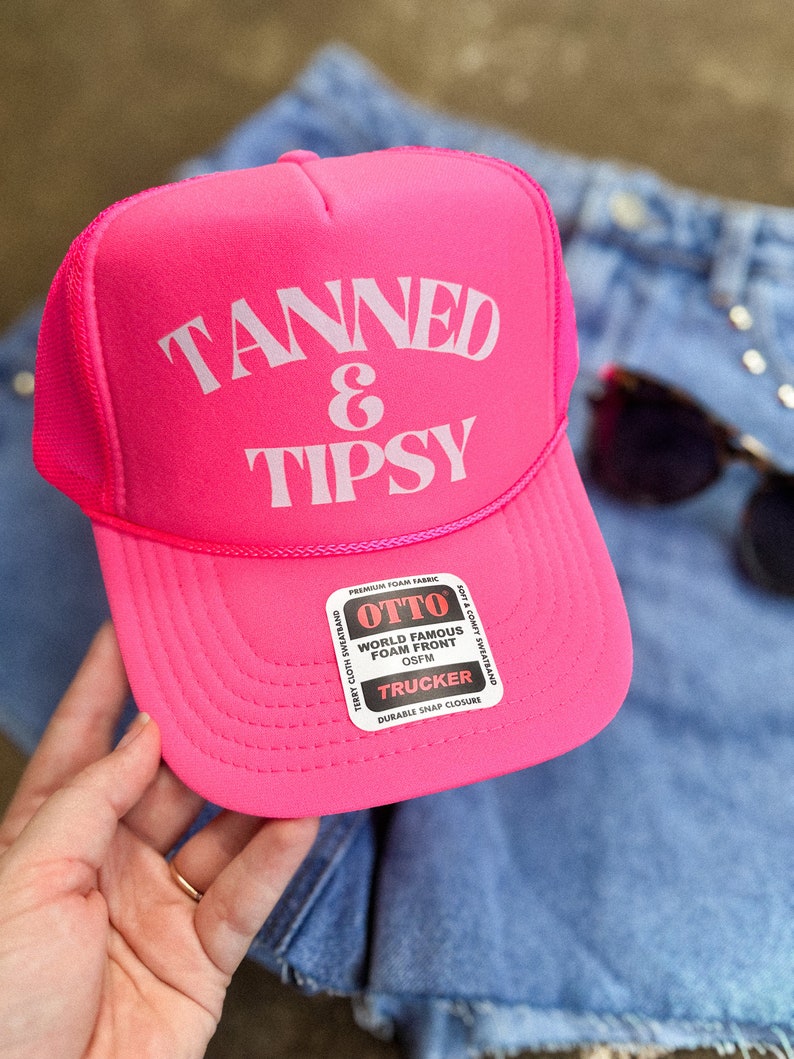 May include: A bright pink trucker hat with the words "TANNED & TIPSY" in white block letters. The hat has a mesh back and a curved brim. The hat also has a tag that says "OTTO" and "TRUCKER".