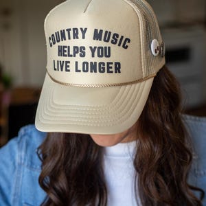 Può includere: Cappellino da camionista beige con la scritta "COUNTRY MUSIC HELPS YOU LIVE LONGER" in nero. Il cappello ha un retro in rete e un dettaglio in corda dorata lungo il bordo. Una piccola spilla è attaccata al lato.