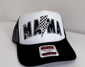 May include: Black and white trucker hat with a white front panel and a black mesh back. The front panel features a black and white checkered lightning bolt design with the word "MAMA" in black letters. The hat has a black snap closure and a black label with the text "OTTO WORLD FAMOUS FOAM FRONTS OSFM TRUCKER DURABLE SNAP CLOSURE".