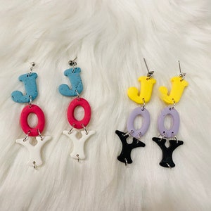 May include: A pair of dangle earrings with the word "JOY" spelled out in colorful letters. The earrings are made of polymer clay and have a silver tone finish.