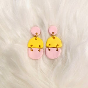 Puede incluir: Un par de pendientes con tres formas geométricas apiladas. La forma superior es un pequeño círculo rosa, la forma del medio es un semicírculo amarillo más grande y la forma inferior es un semicírculo rosa.