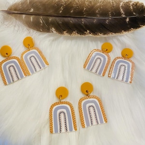 Puede incluir: Un conjunto de cuatro pendientes con temática de arcoíris con un respaldo amarillo mostaza. Los pendientes presentan un fondo blanco con un diseño de arcoíris en tonos grises y negros.