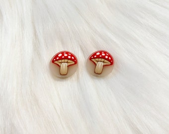 May include: A pair of red and white mushroom stud earrings. The mushrooms have white spots and are on a white background.