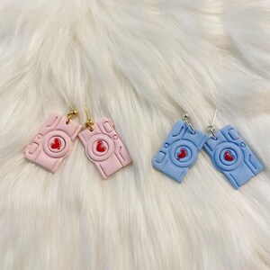 Puede incluir: Dos pares de pendientes con forma de cámara. Un par es de color rosa claro con ganchos dorados, y el otro es azul claro con ganchos plateados. Cada cámara tiene un corazón rojo en el centro.