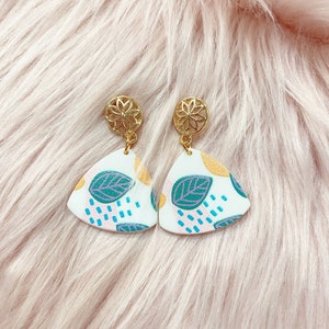 May include: A pair of white triangle-shaped earrings with a gold filigree design at the top. The earrings feature a colorful abstract pattern with green leaves, yellow dots, pink and blue accents.