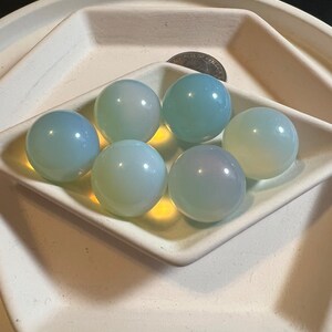 May include: Six smooth, pale blue, round, glass spheres, or orbs, are arranged in a white, diamond-shaped dish. The dish is on top of a larger, white, round dish.