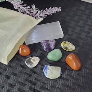 May include: A set of seven tumbled stones in various colors, including clear, red, green, purple, blue, and yellow. The stones are arranged on a black surface with a white selenite stick and a small white pouch.