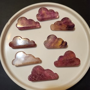 May include: Eight purple, pink, and white fluorite cloud-shaped crystal carvings on a white plate.