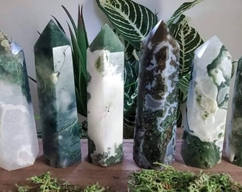 May include: Six green and white agate crystal points, also known as moss agate, are arranged on a wooden surface with green moss in front of them. The crystals are polished and have a natural, organic look.