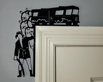 May include: Black metal silhouette wall decor featuring a figure standing next to a van with Christmas lights. The silhouette is mounted on a white door frame.